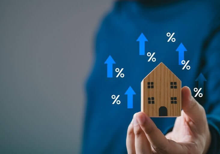 Hand holding wooden house model with upward blue arrows and percentage signs, symbolizing real estate market and mortgage rate increase.